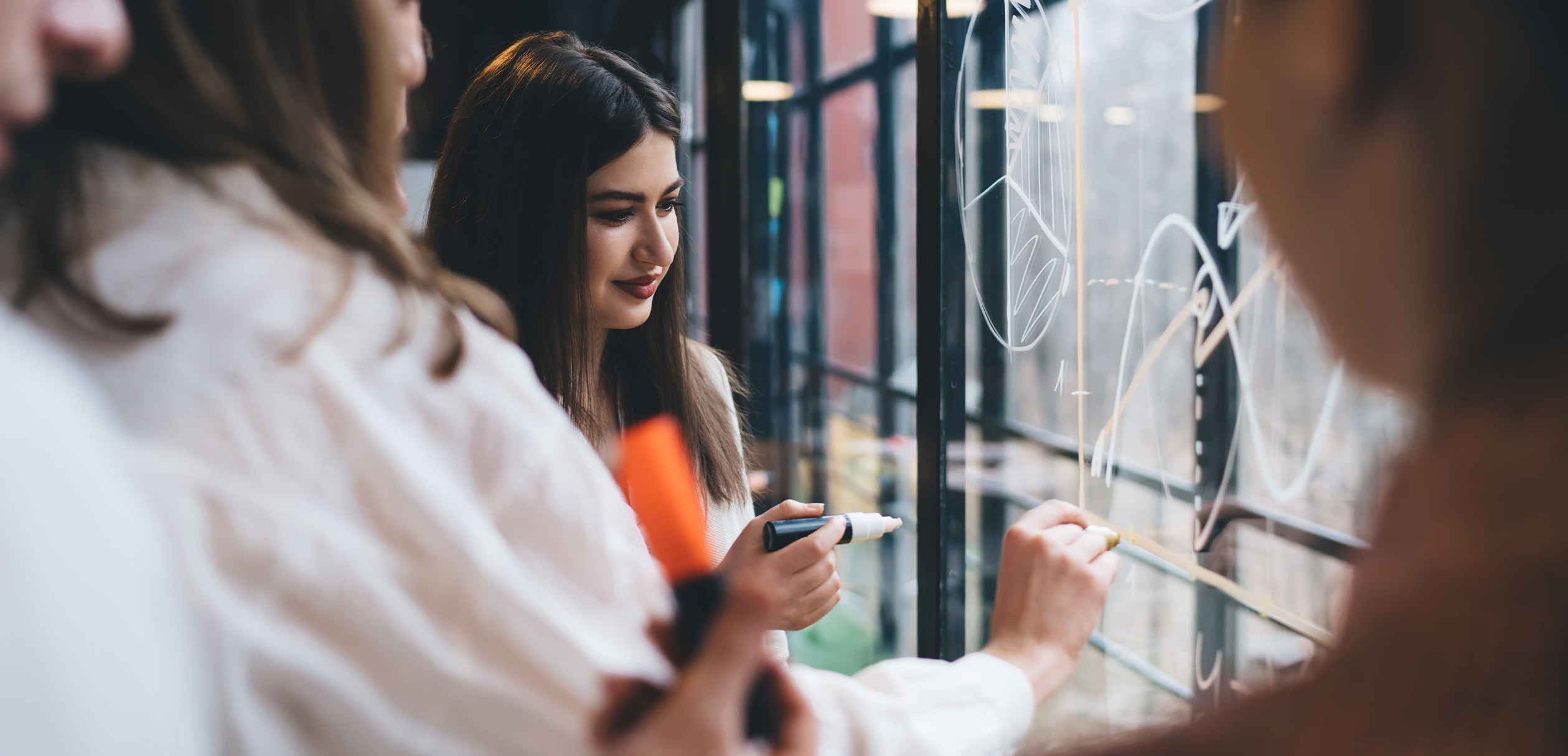 A group of people with markers in hand brainstorming ideas on a clear whiteboard.